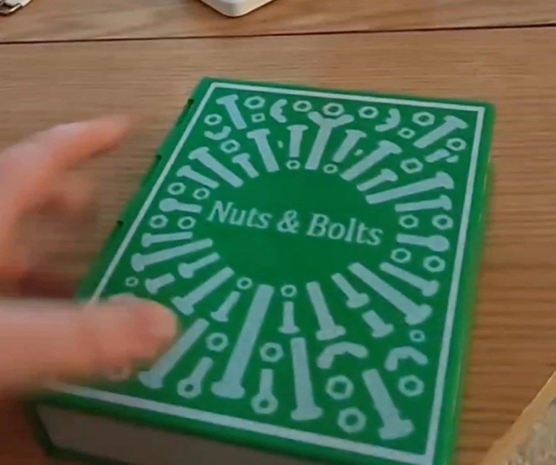 A green 3D-printed book-shaped box on a wooden desk, its cover embossed with an antique 'Nuts & Bolts' design of bolts, nuts and washers radiating around the central title.