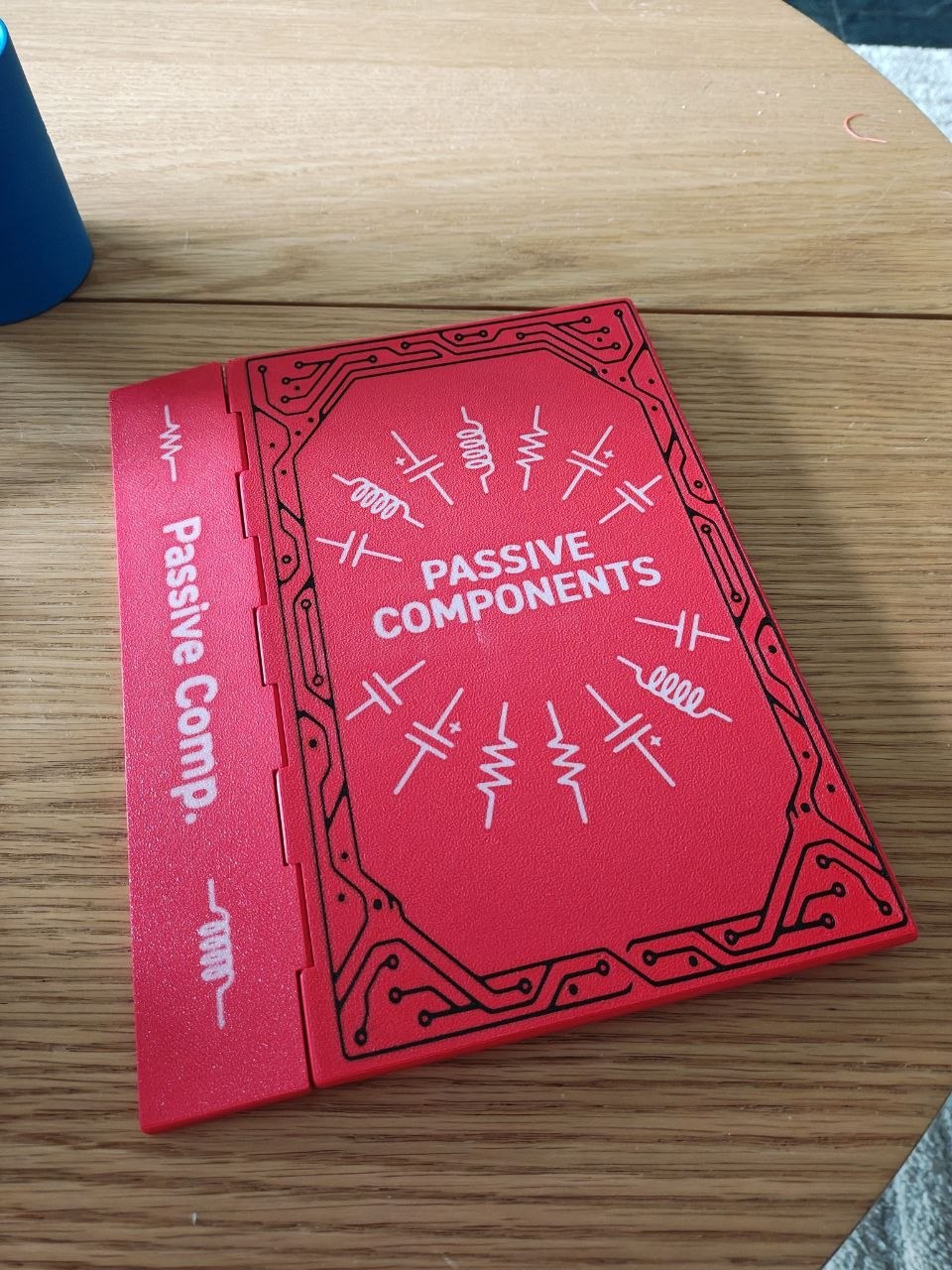 A red 3D-printed book-shaped box on a wooden table, its cover embossed with a circuit-board border and passive-component schematic symbols around the title 'Passive Components', the spine reading 'Passive Comp.'