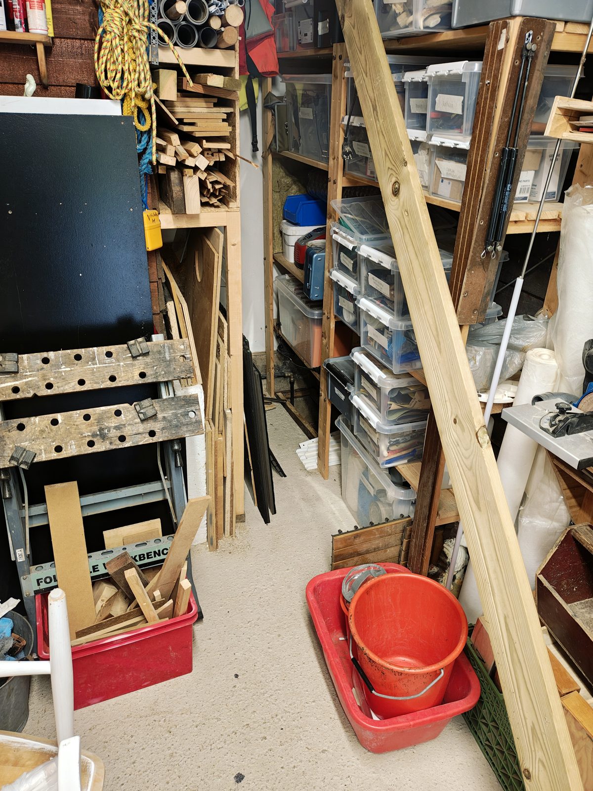 The cluttered corner of a workshop: shelving on both sides packed with plastic bins, wood offcuts and coiled rope, a folding workbench on the left, and a red bucket sitting in a tub on the concrete floor. Boards cover an opening in the floor in the far corner under the shelving.