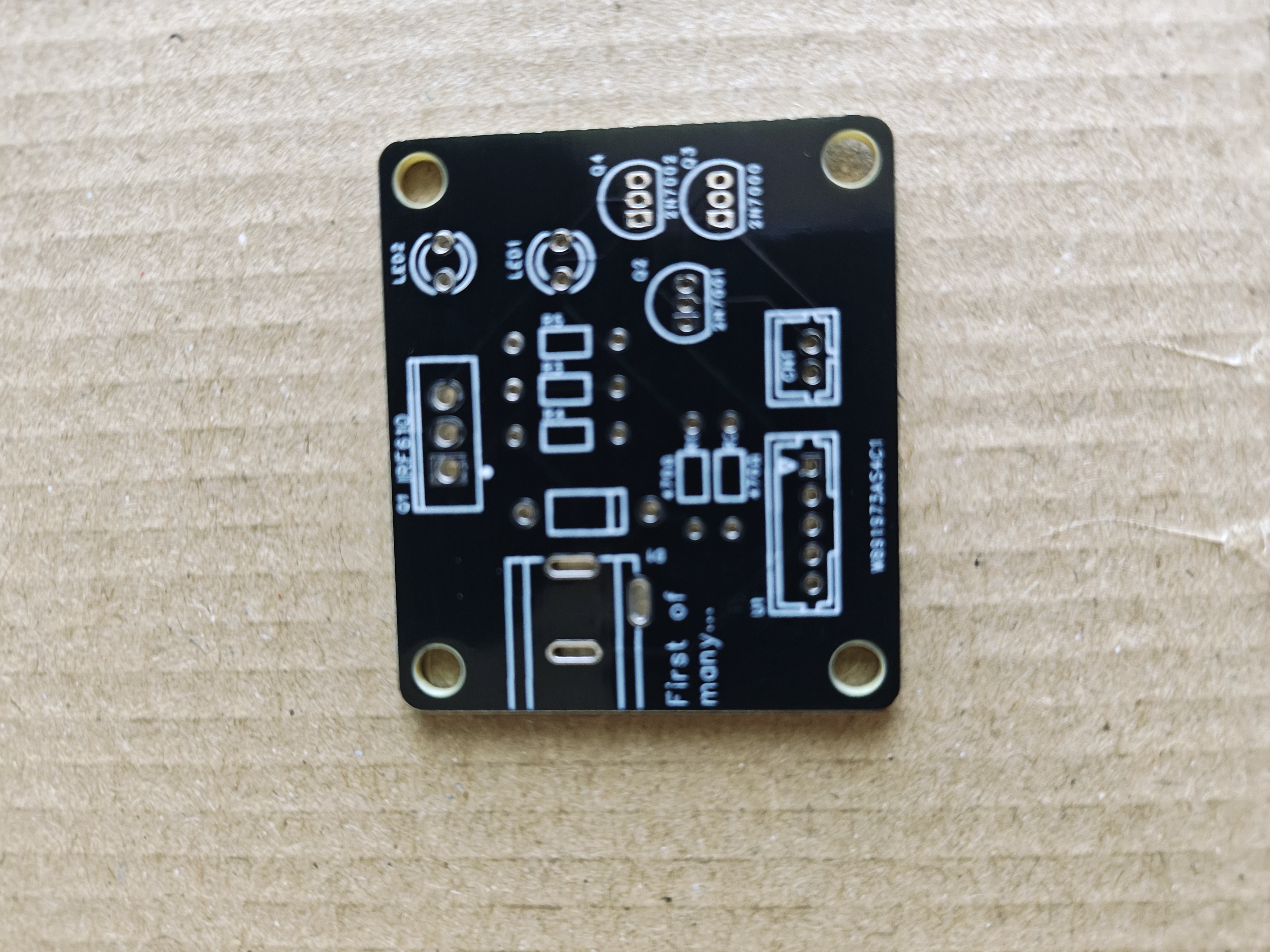 A bare black PCB on a cardboard surface. White silkscreen shows footprints for two LEDs, four transistors, several resistors, two JST connectors and a barrel jack, with 'First of many...' printed in one corner.