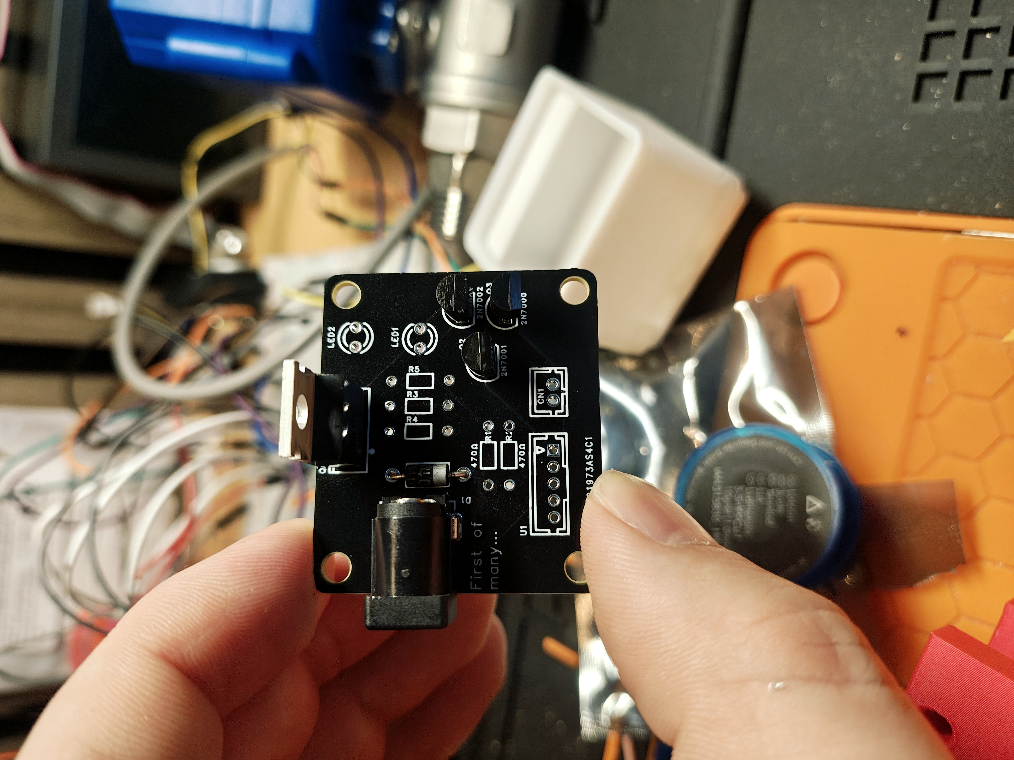 The black board held between two fingers, partly populated. The black barrel jack, the TO-220 IRF610 with its metal tab to the left edge, and the small TO-92 transistors are soldered in; a small axial diode sits in the shadow of the barrel jack. The JST connector and resistor footprints are still empty.