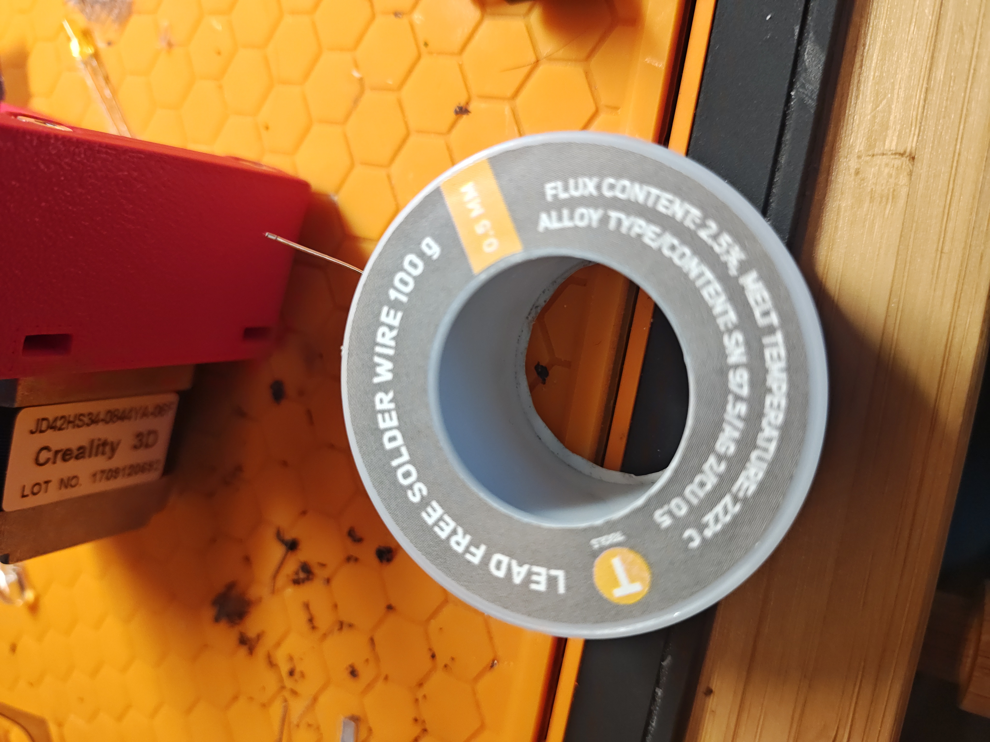 A grey 100 gram reel of lead-free solder wire on an orange honeycomb silicone work mat. The label reads 'LEAD FREE SOLDER WIRE 100 g', 'FLUX CONTENT 2.5%', 'MELT TEMPERATURE 217-227 C', alloy Sn 97.5 / silver 2 / Cu 0.5, 0.5 mm.