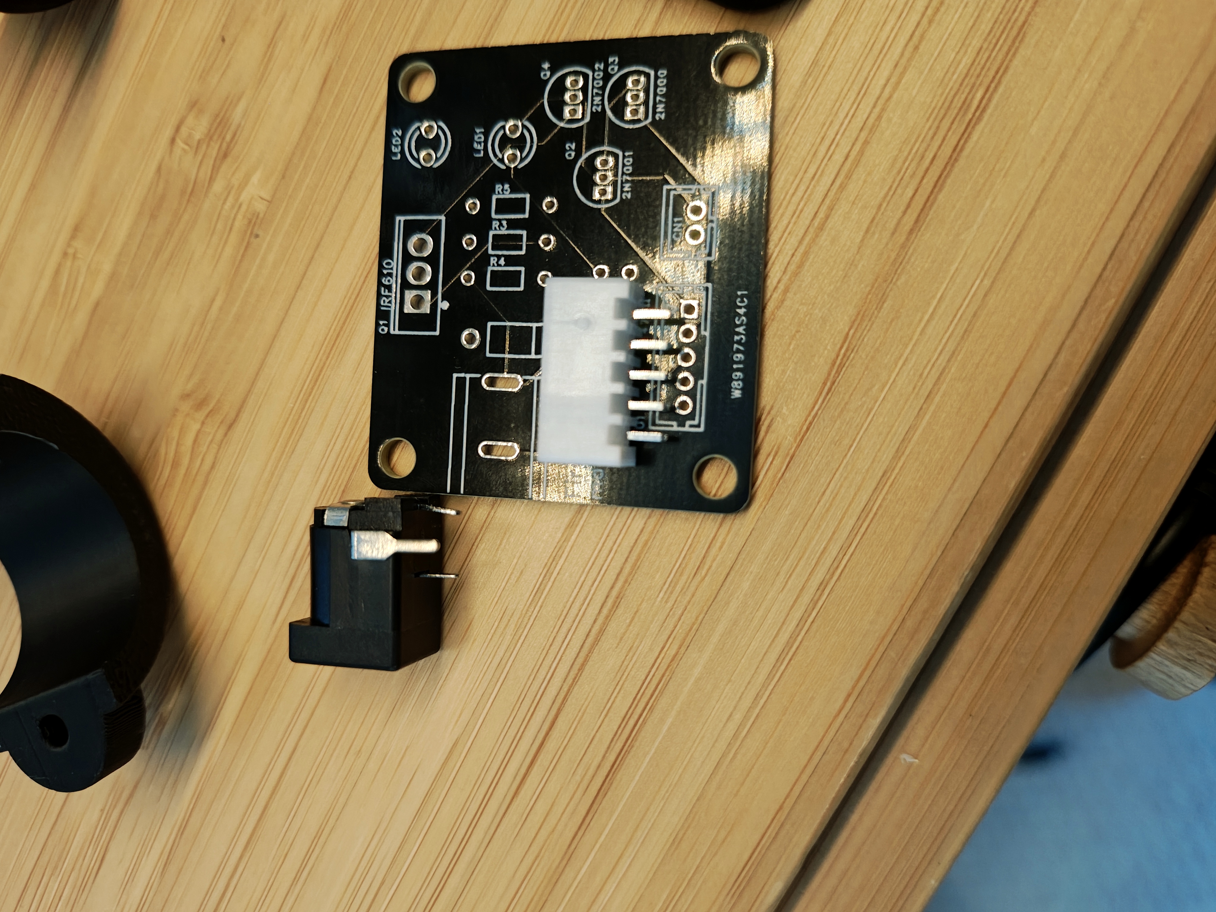 The black board on a wooden bench with a white JST XH connector resting on the connector footprint; its pins sit wider than the holes. A black barrel jack lies loose beside it.